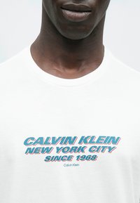 Biały bawełniany T-shirt z okrągłym dekoltem, ozdobiony niebiesko-czerwonym napisem "CALVIN KLEIN NEW YORK CITY SINCE 1968" oraz małym logo.
