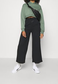 Donna con felpa verde corta, pantaloni neri a gamba larga, scarpe da ginnastica bianche, calzini bianchi e una borsa a tracolla nera su uno sfondo semplice.