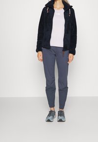 Veste en polaire marine avec col haut et cordons de serrage, t-shirt rose clair, pantalons gris foncé et chaussures de randonnée grises avec des lacets noirs.
