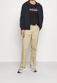 Sudadera con cremallera en color navy con detalles en rosa, camiseta gráfica negra, pantalones beiges y zapatillas beiges. Tejido texturizado, corte estándar, estilo casual.