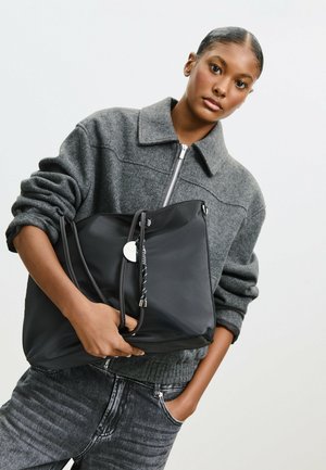 Stor sort tote taske lavet af nylon med snørrelukning, sølv hardware og et mærke. Modellen bærer en grå strikket jakke og denim jeans.