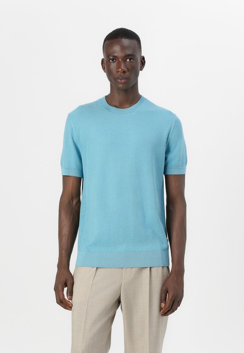 Pulover s kratkimi rokavi, svetlo modre barve, s teksturno vzorcem, okroglim izrezom in rebrastimi manšetami ter robom, v kombinaciji s svetlo bež hlačami.