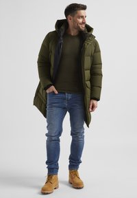 Olivgrüne Daunenjacke mit schwarzem Innenfutter, kombiniert mit einem dunkelgrünen gerippten Pullover, blauen Jeans und braunen Schnürstiefeln.