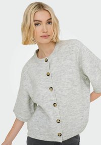 Femme blonde aux cheveux courts portant un cardigan boutonné à manches courtes gris clair sur un pantalon sombre, devant un fond blanc uni.