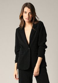 Zwarte blazer gemaakt van een gestructureerde stof met een subtiele glans, voorzien van een enkele knoopsluiting en twee voorzakken, in een getailleerde pasvorm.