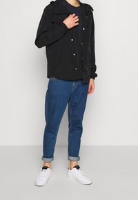 Mies, jolla on musta hupullinen takki, tummansininen paita, käännetyt siniset farkut ja valkoiset tennarit, seisoo yksinkertaista vaaleaa taustaa vasten.