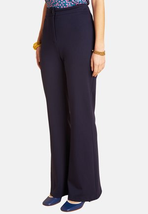 Frau trägt hoch taillierte marineblaue Hosen mit weitem Bein, blaue spitze Schuhe, gemustertes Oberteil und goldene Armbänder an beiden Handgelenken.