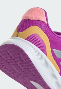 Chaussure de sport violette avec une tige en mesh texturée, languette au talon rose et accents jaunes ; semelle blanche avec un motif pour une adhérence optimale.