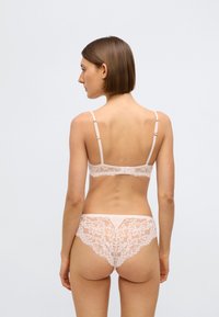 Mežģīņu apakšveļas komplekts maigi rozā krāsā, izceļot smalkas ziedu rakstus, regulējamas siksniņas un plānu jostas vietu atbilstošajām biksītēm.