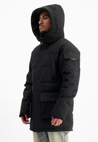 Man in een zwarte winterjas met capuchon en grote zakken, met lichtblauwe jeans tegen een witte achtergrond.