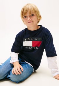 Tricou bleumarin cu logo alb și roșu inscripționat "TOMMY HILFIGER" pe față, asortat cu blugi bleu deschis și mâneci lungi albe pe dedesubt.