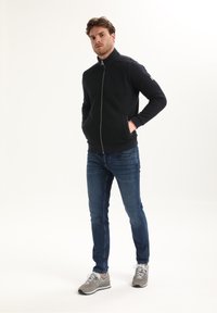 Hombre de pie con las manos en los bolsillos, vistiendo una chaqueta negra con cremallera, jeans azules y zapatillas grises frente a un fondo sencillo.