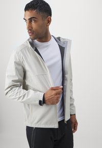 Giacca grigia chiara con cappuccio, dotata di chiusura con zip, fodera in rete e accenti neri a contrasto. Indossata sopra una camicia bianca.