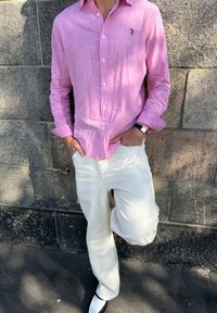 Personne appuyée contre un mur en pierre, portant une chemise rose clair à boutons, un pantalon écru, une montre-bracelet et des chaussures blanches.
