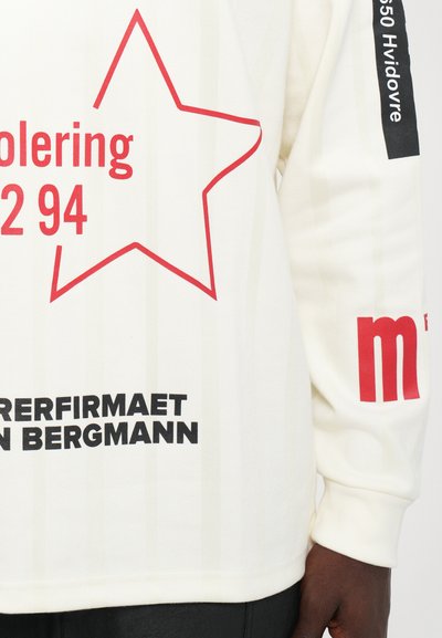 White long-sleeve shirt with red star outline and red and black text, including "Hvidovre" on black strip on sleeve, worn by dark-skinned person.