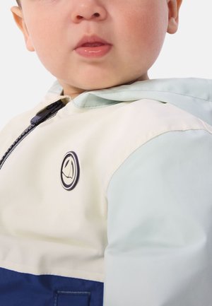 Lichtgekleurde jas met een mintgroene capuchon, een crèmekleurig lichaam en een donkerblauwe onderkant. Voorzien van een rond logo op de borst en een ritssluiting.