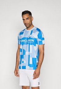 Svetlo modra nogometna majica z geometrijskimi vzorci, kratkimi rokavi, okroglo ovratnico in bele hlače. Na sprednji strani je napis "KONEC".