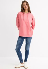 Sudadera rosa con capucha, bolsillo delantero, corte relajado y cordones, combinada con jeans ajustados azules y zapatillas blancas sobre un fondo claro.