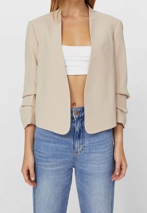 Beiger Blazer mit offenem Frontdesign und gerafften Ärmeln, getragen über einem weißen Crop-Top und hochtaillierten hellblauen Jeans.