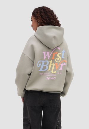 Kvinde med krøllet hår iført en lysegrå hættetrøje med farverig "Wrst Bhvr" tekst og sorte cargobukser, vendt mod venstre foran en ensfarvet baggrund.