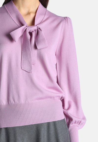 Femme portant un pull à manches longues violet clair avec un nœud noué au niveau de l'encolure et des poignets et ourlet côtelés, associé à une jupe grise.