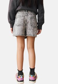 Una persona che indossa pantaloni corti in denim acido, calze nere luccicanti e sneaker colorate, si trova di spalle contro uno sfondo semplice.