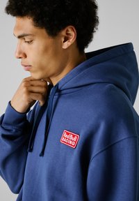 Sudadera azul marino hecha de un tejido suave, con un logo bordado en rojo de "Red Bull RACING" en el pecho. Incluye capucha con cordón y puños acanalados.