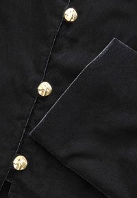 Schwarzer Stoff mit glatter Textur, der goldene dekorative Knöpfe und einen gefalteten Rand aufweist. Das Design beinhaltet Details zur Knopfverschluss.