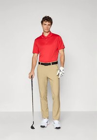 Muž stojí a drží golfovou hůl, má na sobě červené polotričko, béžové kalhoty, bílou golfovou rukavici a bílé tenisky, na jednobarevném šedém pozadí.