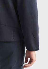 Tamno mornarica flis jakna glatke teksture. Sadrži logo blizu rubova, ravne šavove i detalje od rebraste tkanine na manžetama.