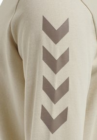 Tissu beige avec quatre flèches chevrons pointant vers le bas, de couleur gris foncé, alignées verticalement sur une manche ou une jambe de pantalon.