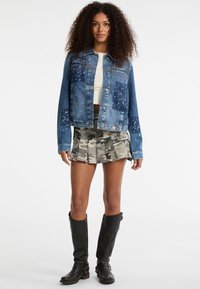 Giacca di denim blu con una vestibilità ampia, dettagli invecchiati e toppe. Abbinata a una gonna plissettata in grigio camouflage e stivali neri fino al ginocchio.