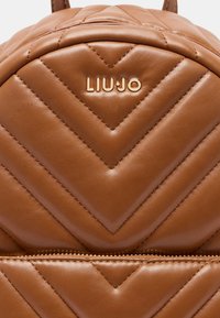 Zaino marrone trapuntato in pelle liscia con motivo a chevron e dettagli del logo dorati, con chiusura a cerniera nella parte inferiore.