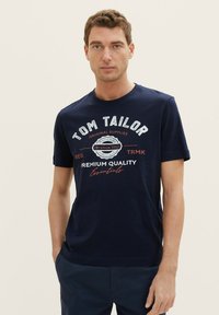 Tamno plavi pamučni majica s okruglim izrezom, kratkim rukavima i ispisanim bijelo/crvenim logotipom s natpisom "Tom Tailor" i dekorativnim elementima.