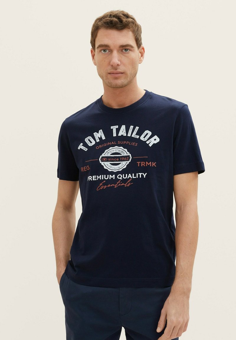 Tamno plavi pamučni majica s okruglim izrezom, kratkim rukavima i ispisanim bijelo/crvenim logotipom s natpisom "Tom Tailor" i dekorativnim elementima.