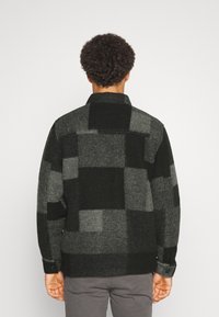 Hombre con chaqueta de lana a cuadros en tonos gris oscuro y negro, visto desde atrás, de pie contra un fondo claro y liso.