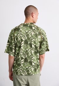 Majica kratkih rukavov v zeleni barvi z abstraktnim rastlinskim vzorcem, ki ima teksturirano tkanino in kontrastne rjave šive ob ovratniku.