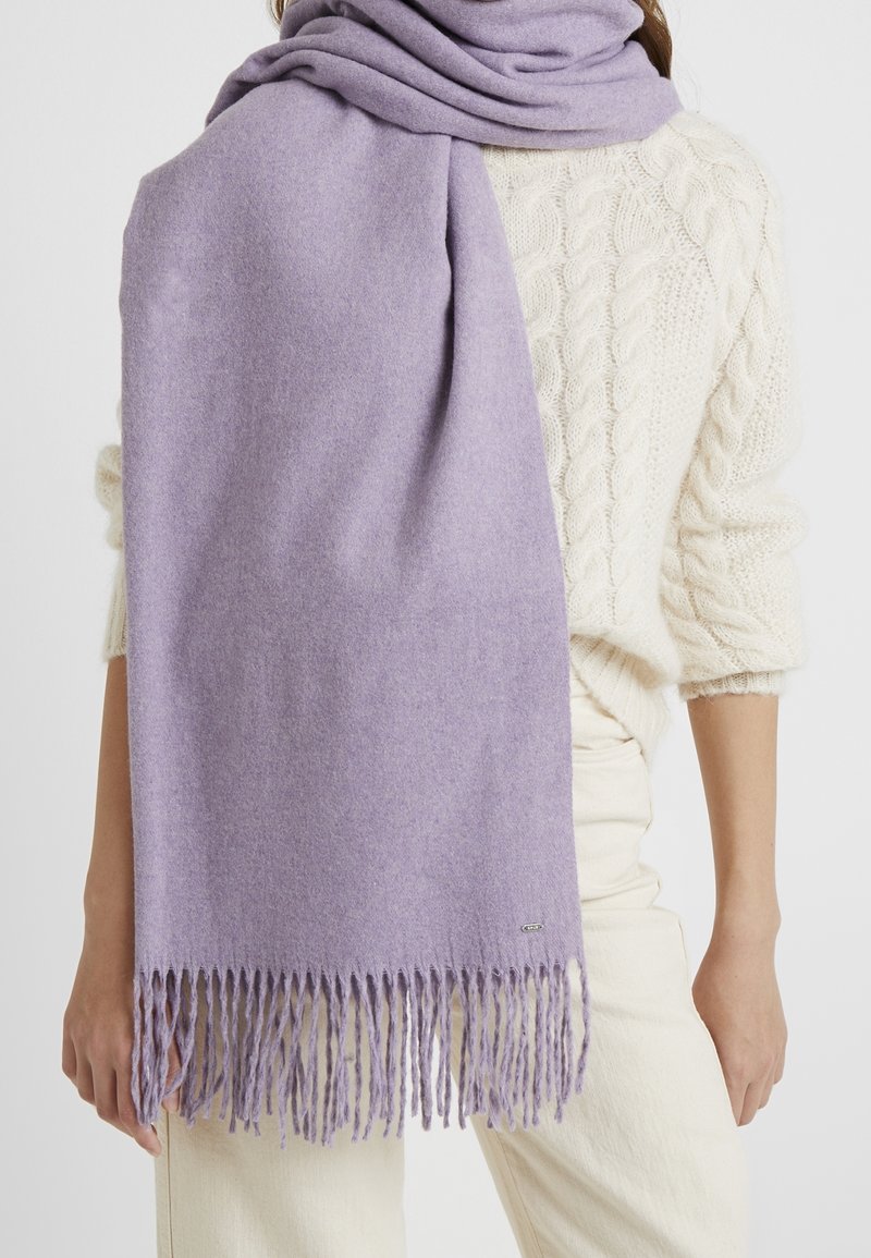 Écharpe lilas à franges drapée sur l’épaule d’une personne portant un pull torsadé crème et un pantalon blanc cassé sur un fond uni.