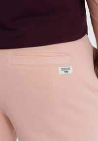 Pantalons de survêtement rose clair avec une poche arrière ornée d'un accent côtelé, une petite étiquette de marque et une finition en tissu doux et texturé.