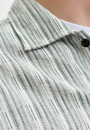 Gros plan d'une chemise à col texturée rayée blanche et grise avec un bouton noir, portée sur un vêtement intérieur blanc uni.