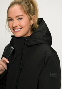 Veste imperméable noire avec un col haut et une capuche réglable, dotée d'une poche poitrine zippée et d'un écusson logo sur la manche.