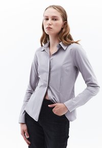 Camisa de manga larga de color gris claro con cuello puntiagudo, cierre de botones y una textura sutil, combinada con jeans oscuros. Diseño simple y ajustado.
