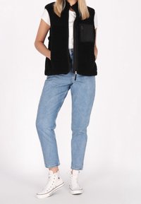 Veste sans manches en polaire noire avec col montant et poche poitrine zippée, associée à un jean denim bleu clair et des baskets montantes blanches.