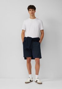 Witte t-shirt, marineblauwe shorts, marineblauwe sweater om de taille geknoopt. Informele schoenen met witte sokken. Eenvoudig ontwerp, effen kleuren, lichtgewicht materiaal.