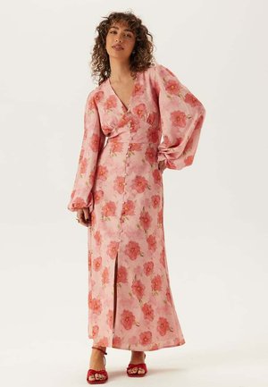 Lång rosa blommig klänning med djup V-ringning, puffärmar, blommönster, sida slits och knapprad fram. Bärs med röda sandaler.