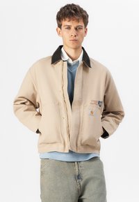 Beige Canvasjacke mit einem dunklen Kragen, vorderem Druckverschluss, zwei großen Taschen und einem kleinen Logo-Patch auf der Brust. Darüber wird ein blauer Pullover getragen.