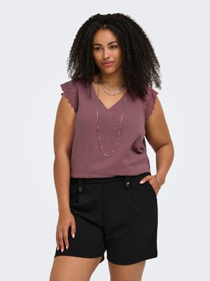 Kvinde med krøllet hår iført en mauve ærmeløs top med blonder i ærmerne og sorte shorts, poserer med den ene hånd i lommen og smiler let.