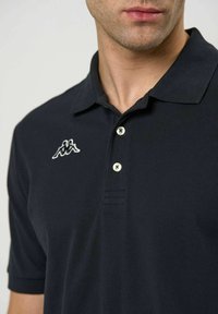 Mann iført en svart kortermet polo-skjorte med en liten hvit logo på venstre bryst og to hvite knapper på stolpen.