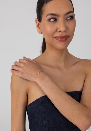 Frau in einem trägerlosen dunkelblauen Oberteil trägt ein filigranes goldenes Armband mit einem kleinen rosa Blumenanhänger und schaut mit einem subtilen Lächeln zur Seite.