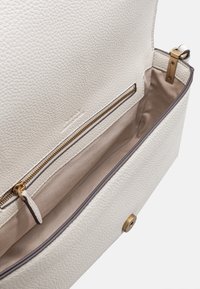 Interno di una borsa a mano in pelle bianca testurizzata, dotato di una tasca con zip e hardware dorato, e foderato in tessuto beige. Chiusura magnetica visibile.